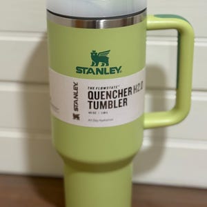 K&ouml;nnte beinhalten: Ein hellgr&uuml;ner Stanley Quencher H2.0 Becher mit Griff. Der Becher hat einen wei&szlig;en und gr&uuml;nen Deckel und ein wei&szlig;es Etikett mit schwarzem Text. Der Becher hat ein Fassungsverm&ouml;gen von 1,18 L und ist f&uuml;r die ganzt&auml;gige Fl&uuml;ssigkeitszufuhr konzipiert.