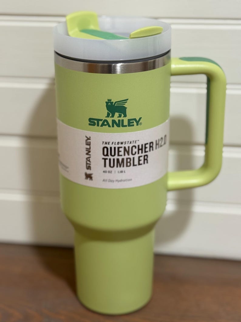 K&ouml;nnte beinhalten: Ein hellgr&uuml;ner Stanley Quencher H2.0 Becher mit Griff. Der Becher hat einen wei&szlig;en und gr&uuml;nen Deckel und ein wei&szlig;es Etikett mit schwarzem Text. Der Becher hat ein Fassungsverm&ouml;gen von 1,18 L und ist f&uuml;r die ganzt&auml;gige Fl&uuml;ssigkeitszufuhr konzipiert.