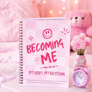 May include: A pink journal with the text "BECOMING ME" and "MY BODY. MY RHYTHM". A pink alarm clock, a pink teddy bear, and a vase of pink flowers are also visible. The journal is designed to help girls grow with confidence, care, and self-respect.