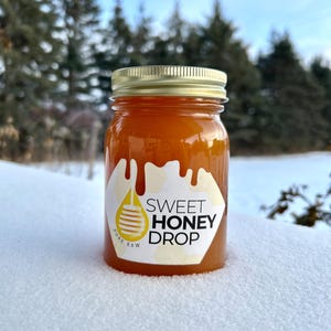 May include: A glass jar filled with golden honey, topped with a gold lid. The jar has a label that reads "Sweet Honey Drop" with a dripping honey design. The background is a snowy landscape with trees.