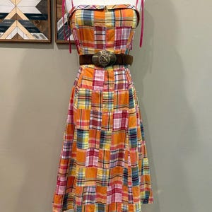 May include: A sleeveless midi dress featuring a patchwork plaid pattern in shades of orange, pink, yellow, and blue. The dress has thin pink shoulder straps and a dark brown suede belt with a large, round, metallic buckle at the waist.