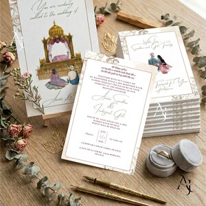 An assortment of wedding stationery cards rests on a wooden surface with two gold fountain pens, ring velvet boxes, and dried floral accents.