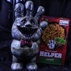 May include: A grotesque, hand-shaped sculpture with a menacing face and a red bow tie stands next to a box of Hamburger Helper Lasagne. The sculpture is a muted green color with stitched details, and the box is red with a picture of the meal.