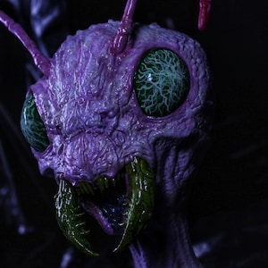 Pode incluir: Close-up de uma criatura insectóide roxa e grotesca com olhos verdes grandes e mandíbulas verdes afiadas. A criatura tem uma aparência texturizada e alienígena, com antenas e uma expressão ameaçadora.