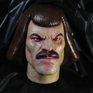Peut inclure: Masque détaillé d'un personnage au visage sévère, avec des cheveux bruns, une moustache foncée et des yeux intenses. Un œil est rouge, l'autre vert. Le masque a un teint pâle et se détache sur un fond sombre.