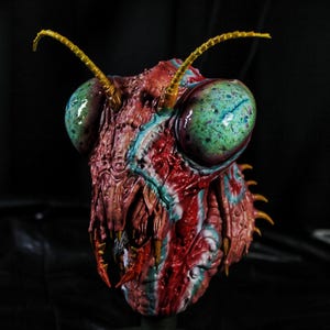 May include: A detailed sculpture of an insect head featuring large, glossy, mottled green compound eyes and a textured, reddish-brown exoskeleton with blue highlights. The creature has two long, segmented yellow antennae and sharp, pointed mandibles.