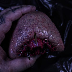 May include: A gruesome, fleshy sculpture with a textured surface and a deep, bloody cavity. The sculpture features multiple black, pointed appendages and a central, sharp element. Held in a hand with tattoos.