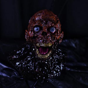 May include: A gruesome, detailed sculpture of a zombie head with exposed skull and decaying flesh. The head has bright blue eyes, an open mouth, and a dark, textured base. The overall color palette is a mix of reds, browns, and blacks, creating a spooky, horror-themed aesthetic.