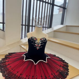 Może przedstawiać: Czarna i czerwona tutu baletowa ze srebrną koroną na manekinie. Gorset jest z czarnego aksamitu ze zdobieniami w kolorze czerwonym i srebrnym. Spódnica jest z warstwowego czerwonego tiulu z czarnym koronkowym wykończeniem i perłowymi akcentami. Kostium baletowy.