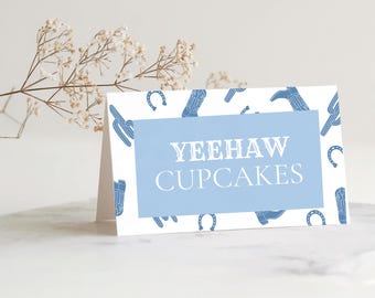 Könnte beinhalten: Eine gefaltete weiße Karte mit einem hellblauen Rechteck in der Mitte mit dem weißen Text "YEEHAW CUPCAKES". Die Karte hat ein Muster aus blauen Cowboy-Stiefeln, Hufeisen und Kakteen. Ein Zweig weißer Blumen befindet sich im Hintergrund.