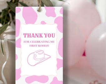 Könnte beinhalten: Ein rechteckiges Geschenk-Etikett mit rosa Kuhmuster-Hintergrund. Das Etikett zeigt den Text "THANK YOU FOR CELEBRATING MY FIRST RODEO!" und eine Illustration eines Cowboyhuts. Ein goldener Faden ist zum Aufhängen befestigt.