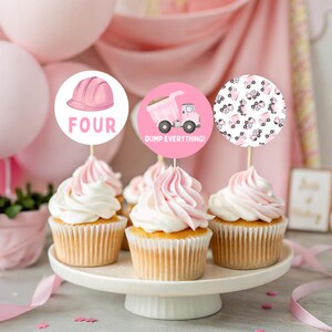 May include: Cupcakes with pink frosting and decorative toppers. One topper features a pink construction hat and the word "FOUR". Another has a pink dump truck and the phrase "DUMP EVERYTHING!". The third has a pattern of pink trucks.
