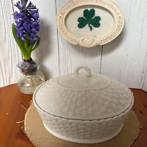 May include: A white ceramic basket-weave covered dish with a looped handle sits on a burlap mat. A framed oval with a green shamrock and the words "Love, Loyalty, Friendship" hangs above. A purple hyacinth flower is in a glass vase.