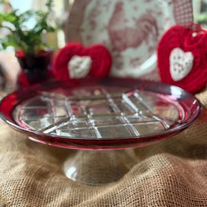 May include: A clear glass cake stand with a red rim and a grid pattern on the surface. Two red heart-shaped decorations with white accents are in the background, along with a decorative plate with a rooster design.