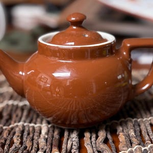 May include: A brown ceramic teapot with a lid and handle. The teapot has a glossy finish and a white rim around the opening. The teapot is sitting on a woven mat.