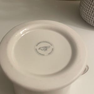May include: A white ceramic mug with a rounded base and a small handle. The bottom of the mug features a circular design with a logo. The mug is set against a white background.