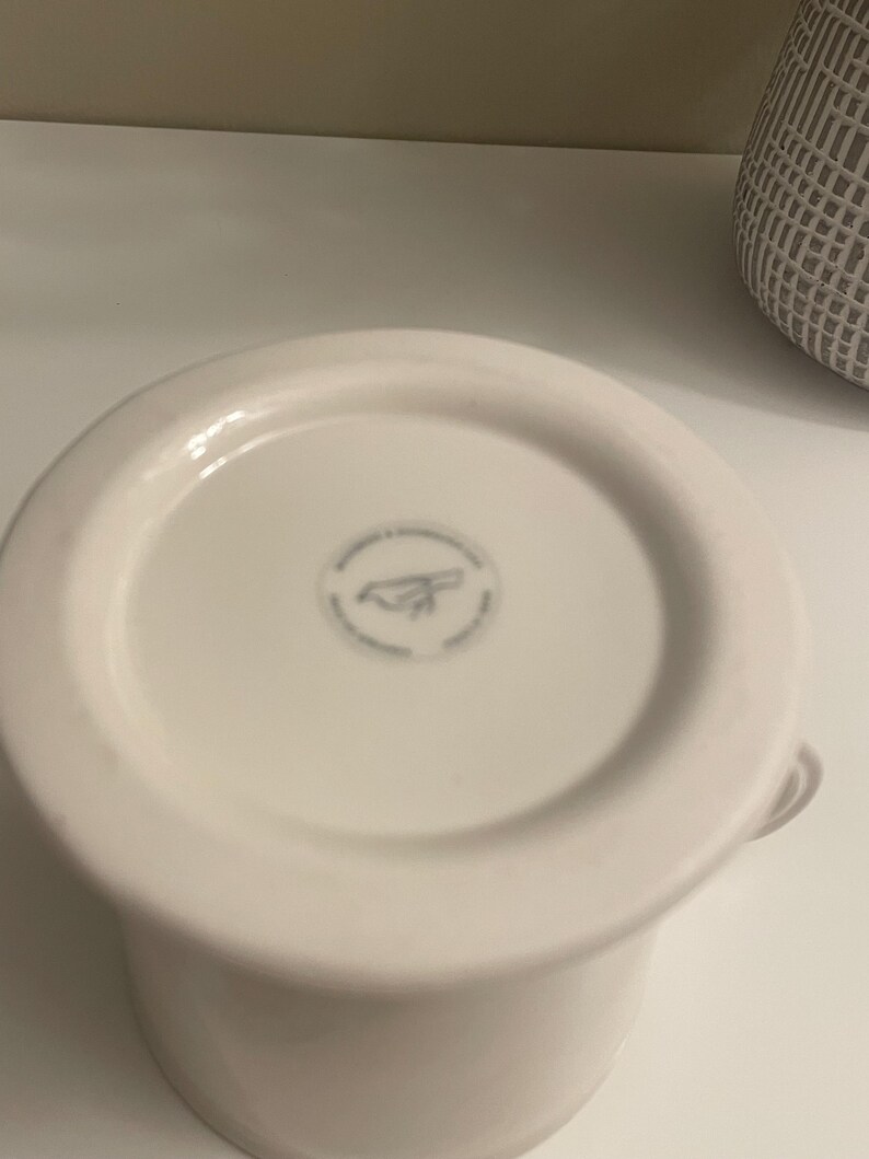 May include: A white ceramic mug with a rounded base and a small handle. The bottom of the mug features a circular design with a logo. The mug is set against a white background.