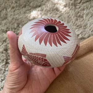 May include: A small, round, off-white ceramic vessel featuring a dark red sunburst pattern around the top opening and a repeating dark red fish motif around the body. The surface has a textured, carved appearance with fine, radiating lines.