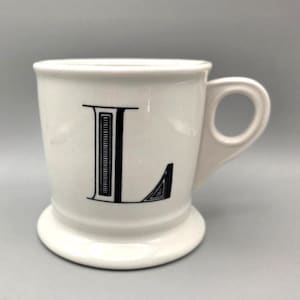 May include: White ceramic mug with a black letter "L" on the front. The mug has a rounded handle and a pedestal base. The letter "L" is in a bold, serif font.