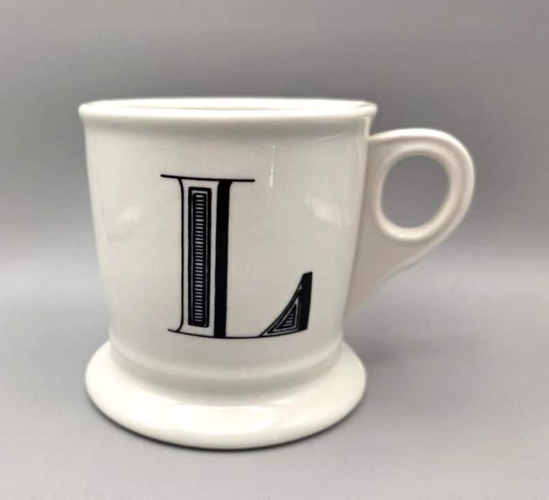 May include: White ceramic mug with a black letter "L" on the front. The mug has a rounded handle and a pedestal base. The letter "L" is in a bold, serif font.