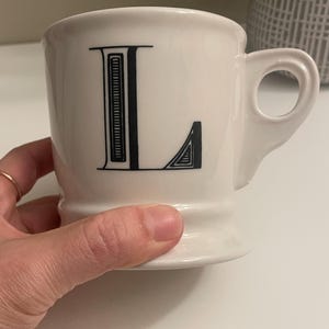 May include: A white ceramic mug with a black letter "L" printed on the front. The mug has a rounded shape, a handle, and a pedestal base. The mug is held by a hand.