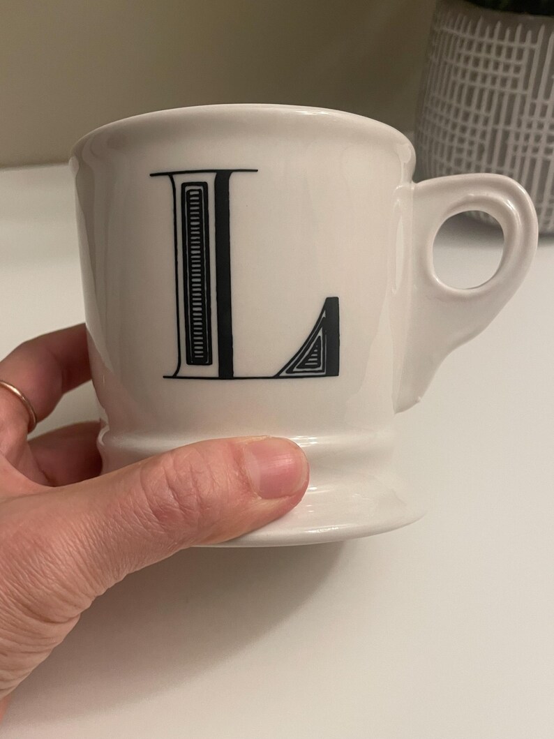 May include: A white ceramic mug with a black letter "L" printed on the front. The mug has a rounded shape, a handle, and a pedestal base. The mug is held by a hand.