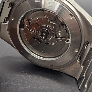 May include: Close-up of the back of a silver wristwatch, showcasing the intricate internal mechanism. The watch has a round, clear back, revealing gears and other components. The watch band is also silver.