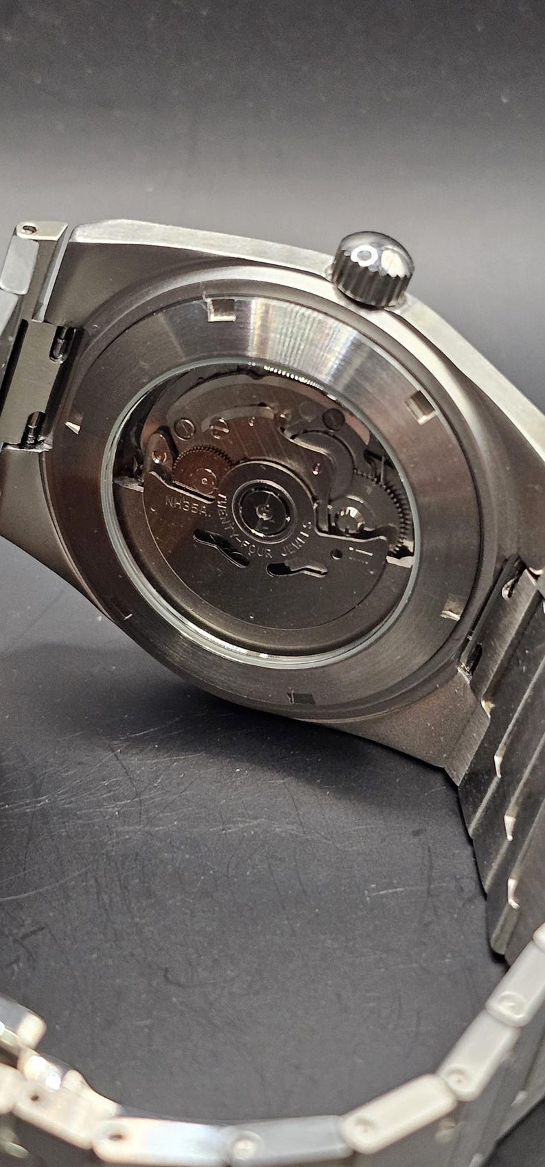 May include: Close-up of the back of a silver wristwatch, showcasing the intricate internal mechanism. The watch has a round, clear back, revealing gears and other components. The watch band is also silver.