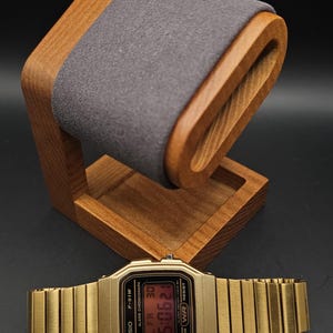 May include: A gold-toned Casio F-91W digital watch with a matching metal band rests below a wooden watch stand with a gray felt cushion. The watch displays the time 5:06:21.