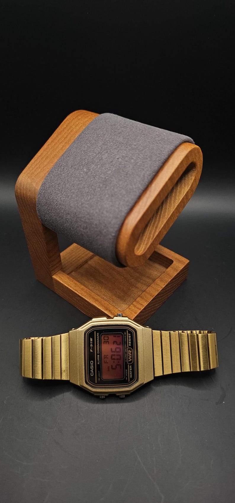 May include: A gold-toned Casio F-91W digital watch with a matching metal band rests below a wooden watch stand with a gray felt cushion. The watch displays the time 5:06:21.