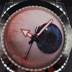 May include: A stainless steel wristwatch featuring a dial designed to resemble a human eye with a blue iris, a black pupil, and red blood vessels. The watch has a silver-toned metal link band, a fluted bezel, and hands with red accents.