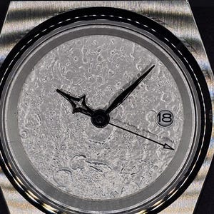 May include: A stainless steel wristwatch with a textured, light gray dial. The watch has black hands and a date window at the 3 o'clock position. The watchband is also stainless steel.