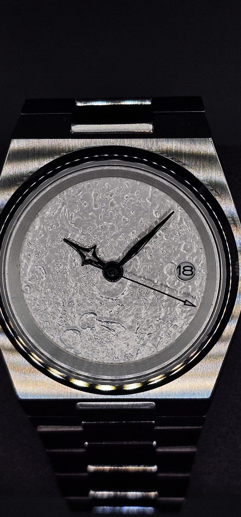 May include: A stainless steel wristwatch with a textured, light gray dial. The watch has black hands and a date window at the 3 o'clock position. The watchband is also stainless steel.