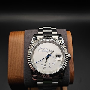 Puede incluir: Un reloj de pulsera plateado con esfera blanca y bisel estriado. El reloj tiene una indicación de fecha a las 3 en punto y el texto "Who cares I'm already late" en la esfera. El reloj se muestra sobre un soporte de madera.