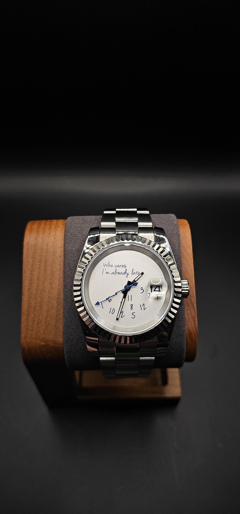 Pu&ograve; includere: Un orologio da polso argentato con quadrante bianco e lunetta scanalata. L'orologio ha un'indicazione della data alle 3 e il testo "Who cares I'm already late" sul quadrante. L'orologio &egrave; esposto su un supporto di legno.