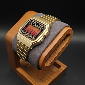 May include: A gold-toned digital watch with a matching band rests on a wooden watch stand. The watch face displays the time in red digits. The stand is made of light brown wood and has a gray cushion.