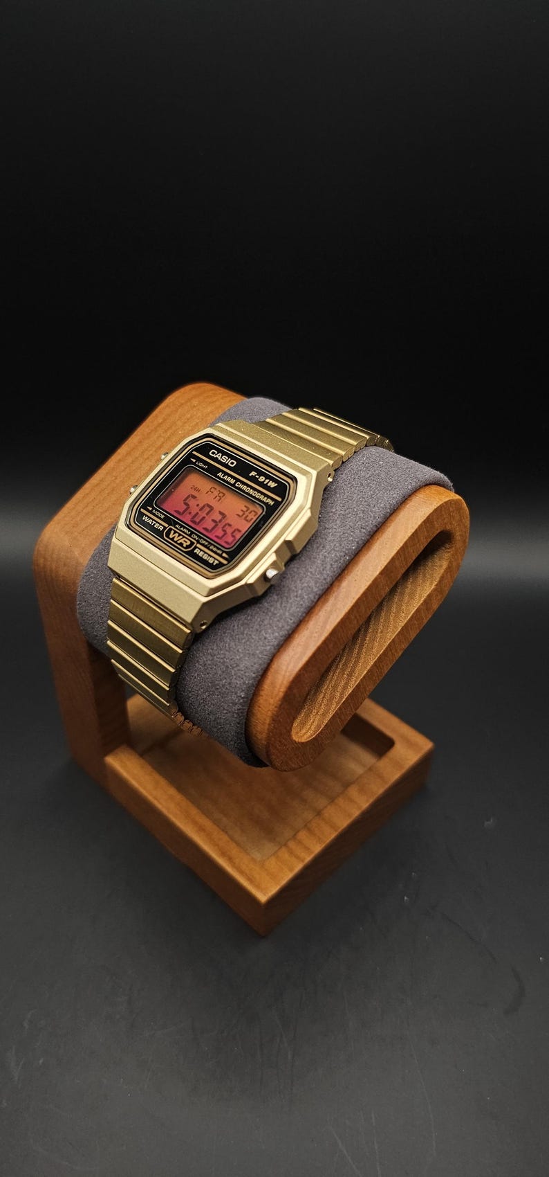 May include: A gold-toned digital watch with a matching band rests on a wooden watch stand. The watch face displays the time in red digits. The stand is made of light brown wood and has a gray cushion.