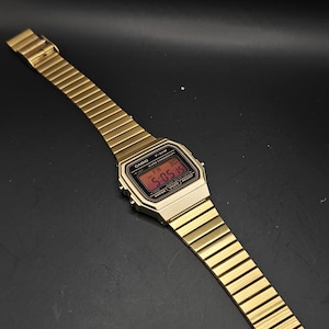 May include: Gold-toned digital watch with a rectangular face and a matching metal band. The watch displays the time in red digits. The watch is a classic style, with a sleek, minimalist design.