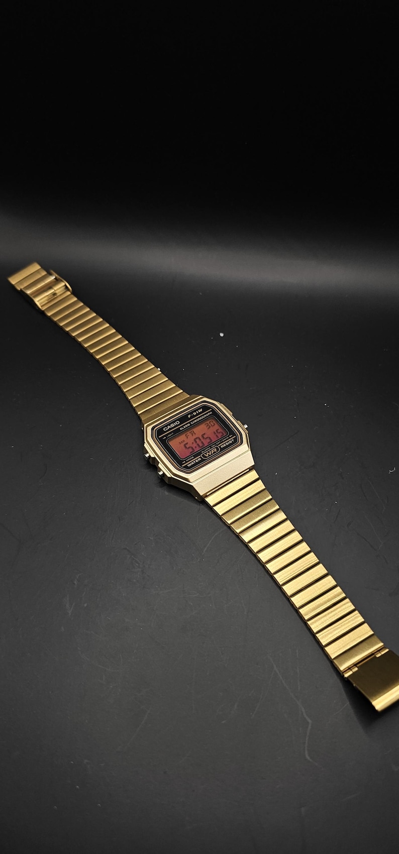 May include: Gold-toned digital watch with a rectangular face and a matching metal band. The watch displays the time in red digits. The watch is a classic style, with a sleek, minimalist design.