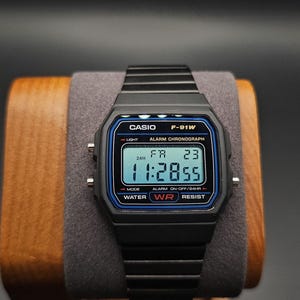 May include: Black Casio F-91W digital watch with a blue border, displaying the time as 11:28:55. The watch is on a wooden and gray display stand. The watch has the text "CASIO F-91W ALARM CHRONOGRAPH" on the face.