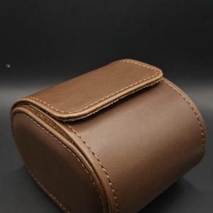May include: A brown leather watch case with a rounded rectangular shape. The case has a flap closure and is stitched with a lighter brown thread. The background is a dark gray.