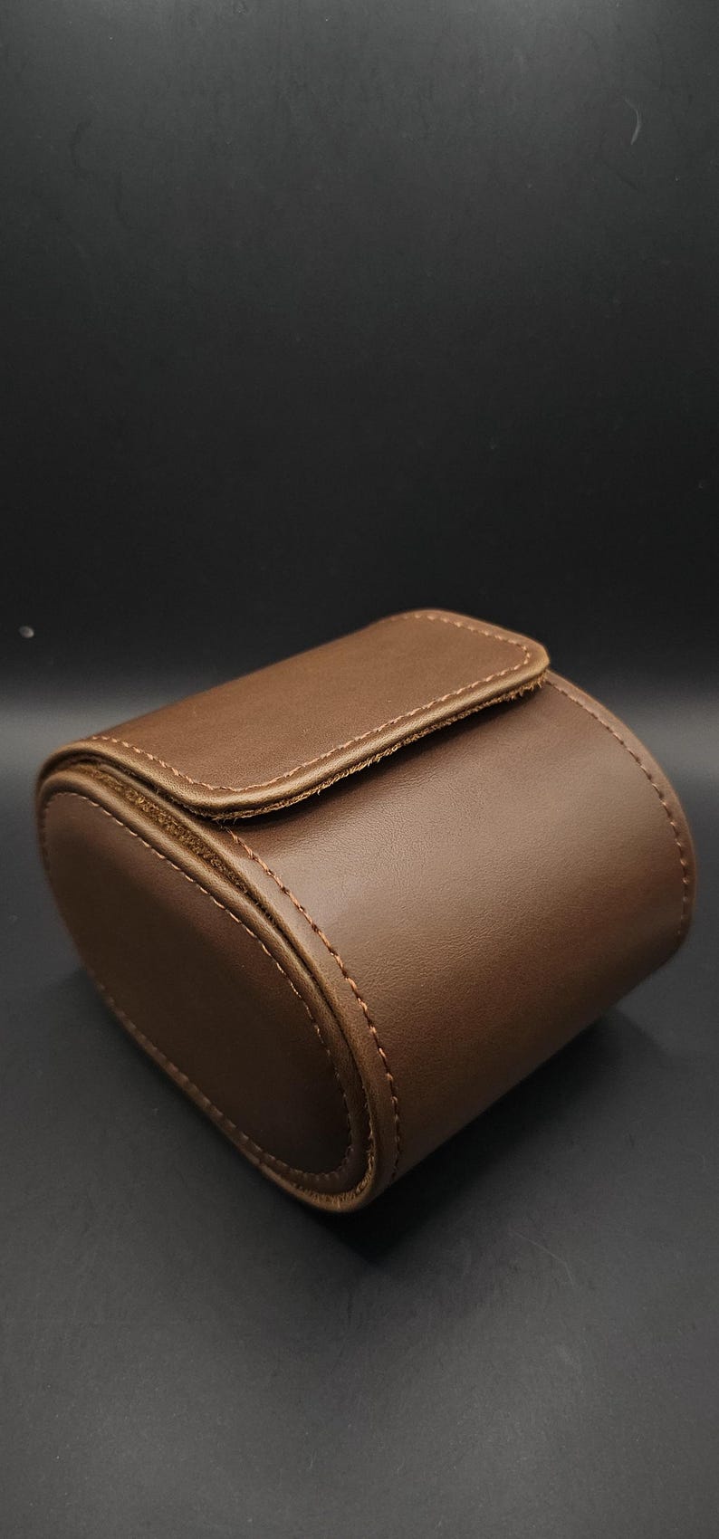 May include: A brown leather watch case with a rounded rectangular shape. The case has a flap closure and is stitched with a lighter brown thread. The background is a dark gray.