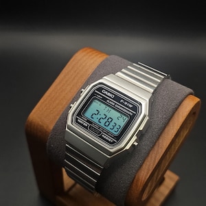 Puede incluir: Un reloj digital Casio F-91W plateado con una pantalla negra que muestra la hora. El reloj está sobre un soporte de madera con un cojín gris. La correa del reloj es plateada, sobre un fondo negro.
