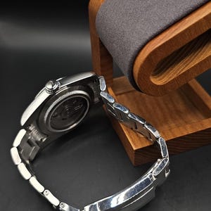 Pu&ograve; includere: Un orologio da polso argentato con cinturino in metallo, appoggiato su un supporto in legno per orologi con un cuscino grigio. L'orologio ha un quadrante rotondo e un retro dettagliato. Il supporto &egrave; in legno chiaro.