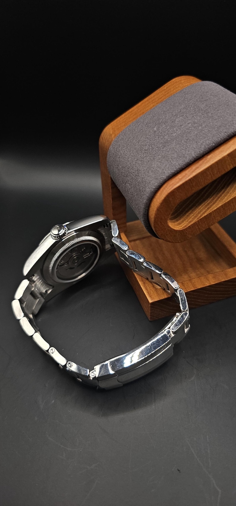 Pu&ograve; includere: Un orologio da polso argentato con cinturino in metallo, appoggiato su un supporto in legno per orologi con un cuscino grigio. L'orologio ha un quadrante rotondo e un retro dettagliato. Il supporto &egrave; in legno chiaro.