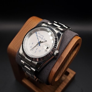 Pu&ograve; includere: Un orologio da polso argentato con quadrante bianco e lunetta scanalata &egrave; appoggiato su un supporto in legno. L'orologio ha un datario e un cinturino in metallo. Sul quadrante c'&egrave; la scritta "Who cares, I'm already late."