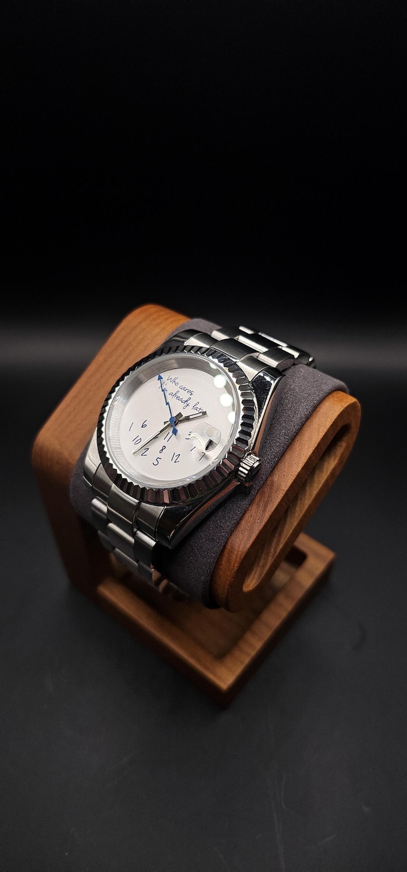 Pu&ograve; includere: Un orologio da polso argentato con quadrante bianco e lunetta scanalata &egrave; appoggiato su un supporto in legno. L'orologio ha un datario e un cinturino in metallo. Sul quadrante c'&egrave; la scritta "Who cares, I'm already late."