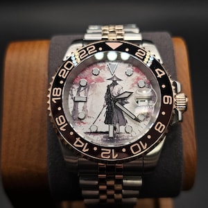 Könnte beinhalten: Luxus-Armbanduhr mit zweifarbigem Edelstahl- und Goldarmband. Das Zifferblatt zeigt eine einzigartige Illustration einer Figur mit Hut vor einem rosa-weißen Hintergrund. Die Lünette hat schwarze und goldene Akzente.