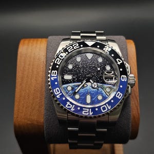 May include: A stainless steel wristwatch with a black and blue rotating bezel. The watch face features a starry night sky design with a blue and white earth graphic. The watch is displayed on a wooden watch holder.