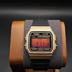 May include: A gold-toned Casio F-91W digital watch with a black strap. The watch face displays the time 1:11:13 and the text "ALARM CHRONOGRAPH". The watch is displayed on a wooden stand.